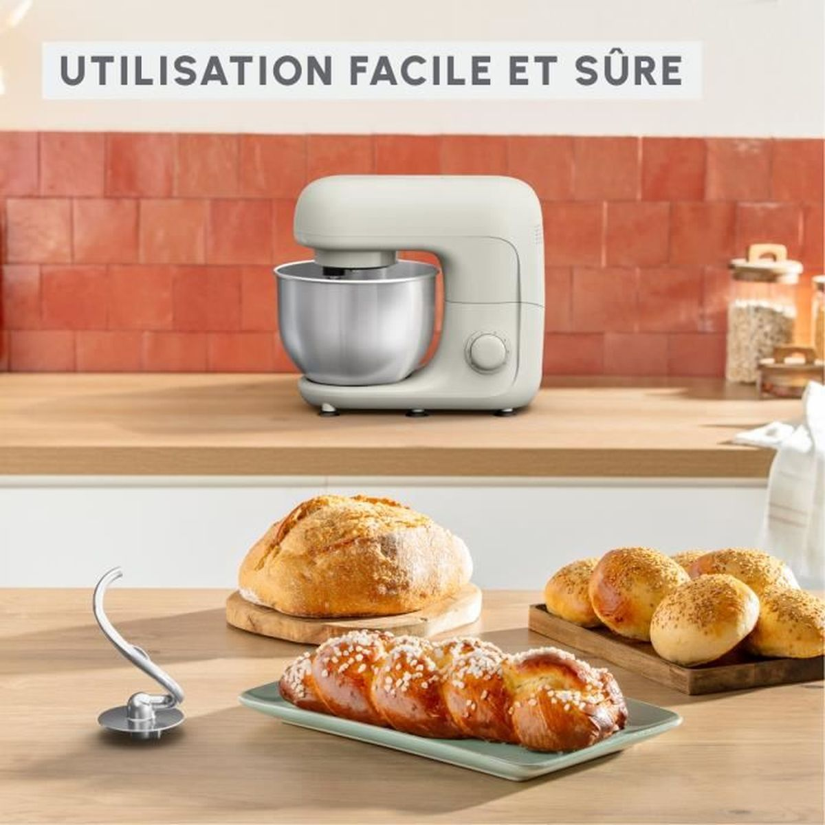 Impastatrice beige su piano di lavoro circondata da pane e dolci davanti a una parete di piastrelle rosse.