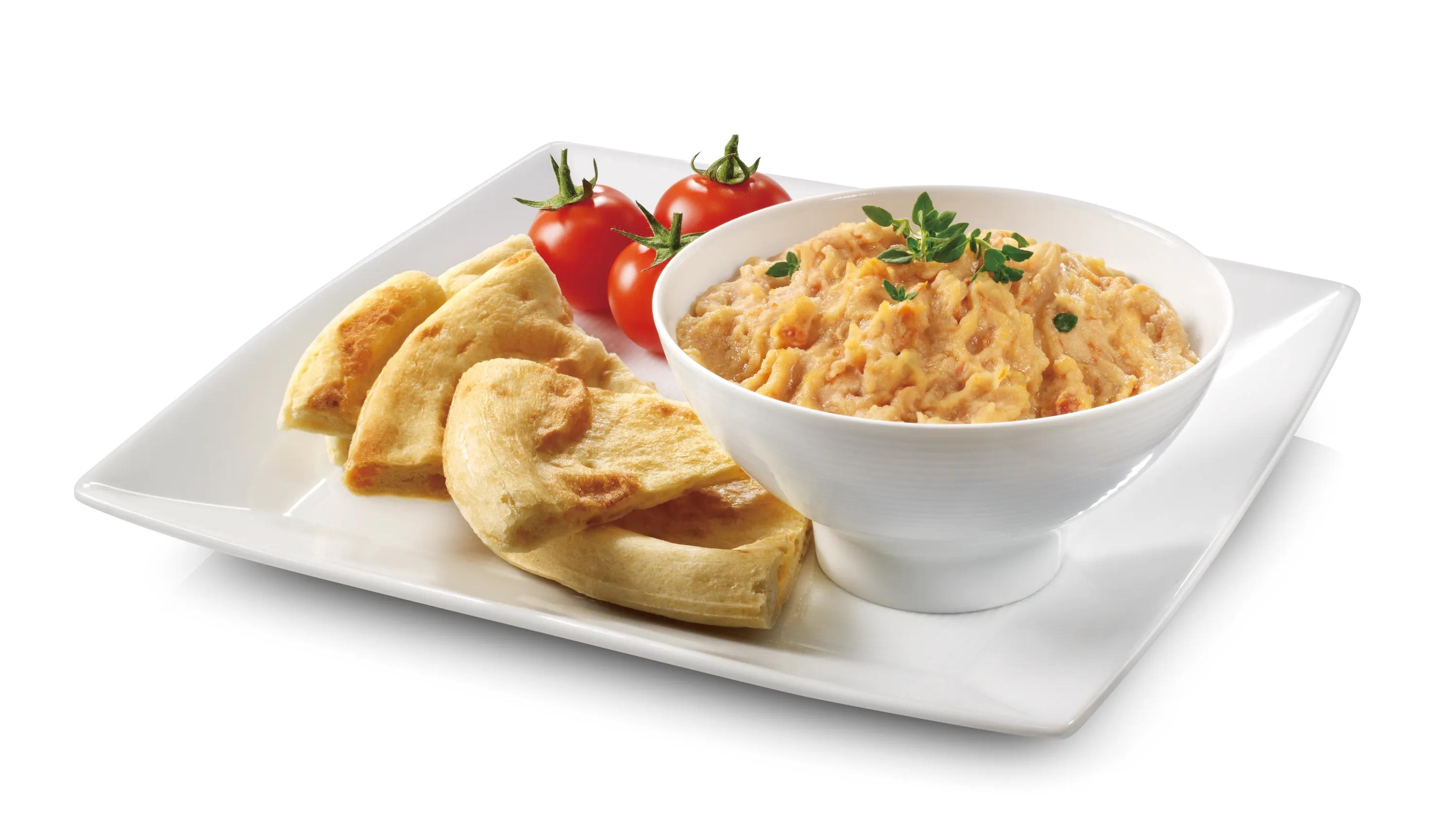 Una ciotola di crema di fagioli con erbe, pane pita e pomodorini su un piatto bianco.
