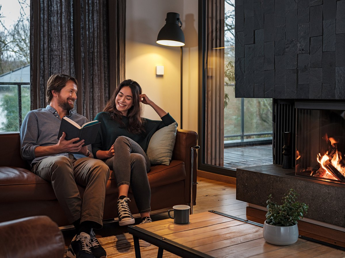 A smiling couple sits on a leather sofa reading a book near a lit fireplace in a cozy, modern living room.