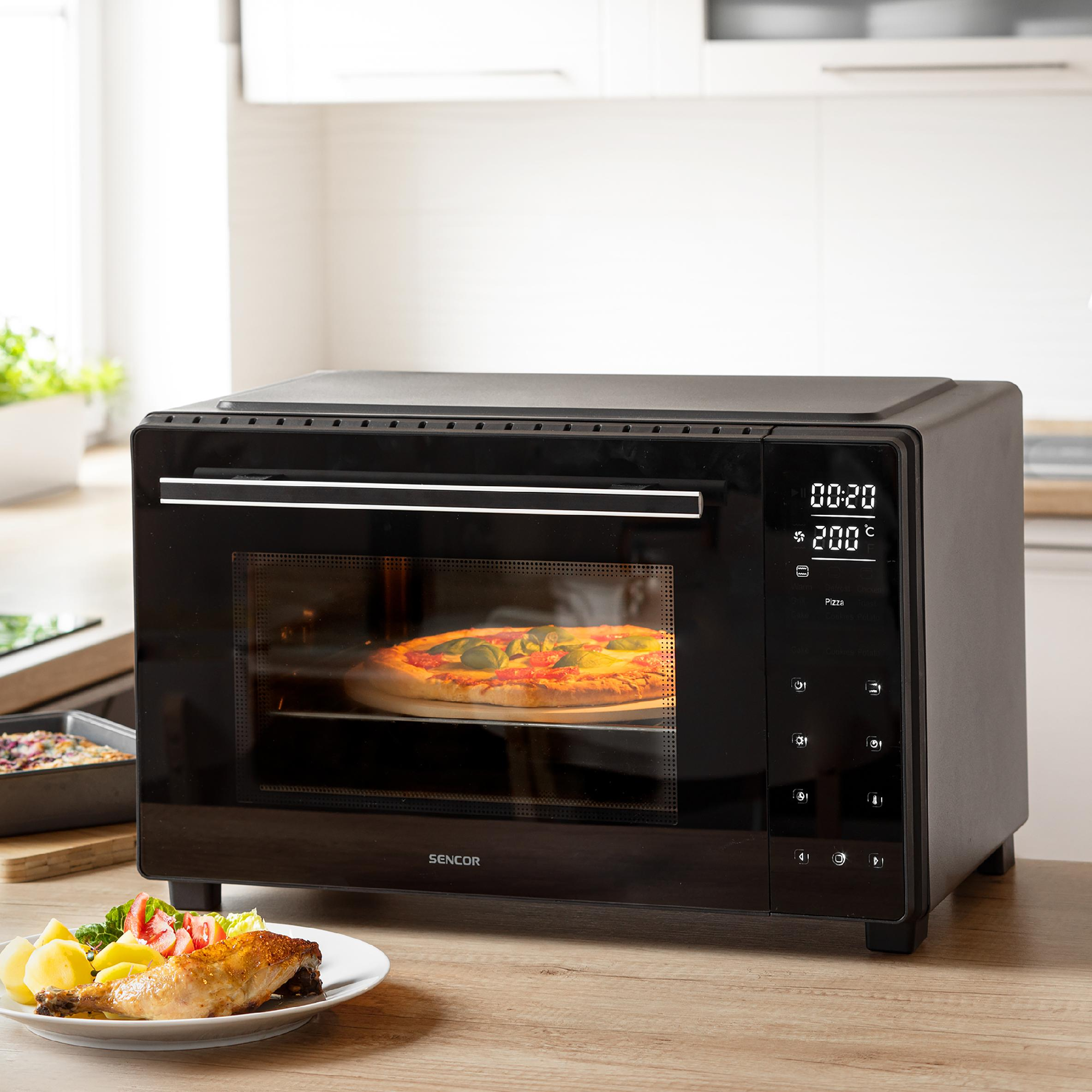 Un mini forno Sencor nero su un piano di lavoro in legno cuoce una pizza, con un piatto di pollo e patate in primo piano.