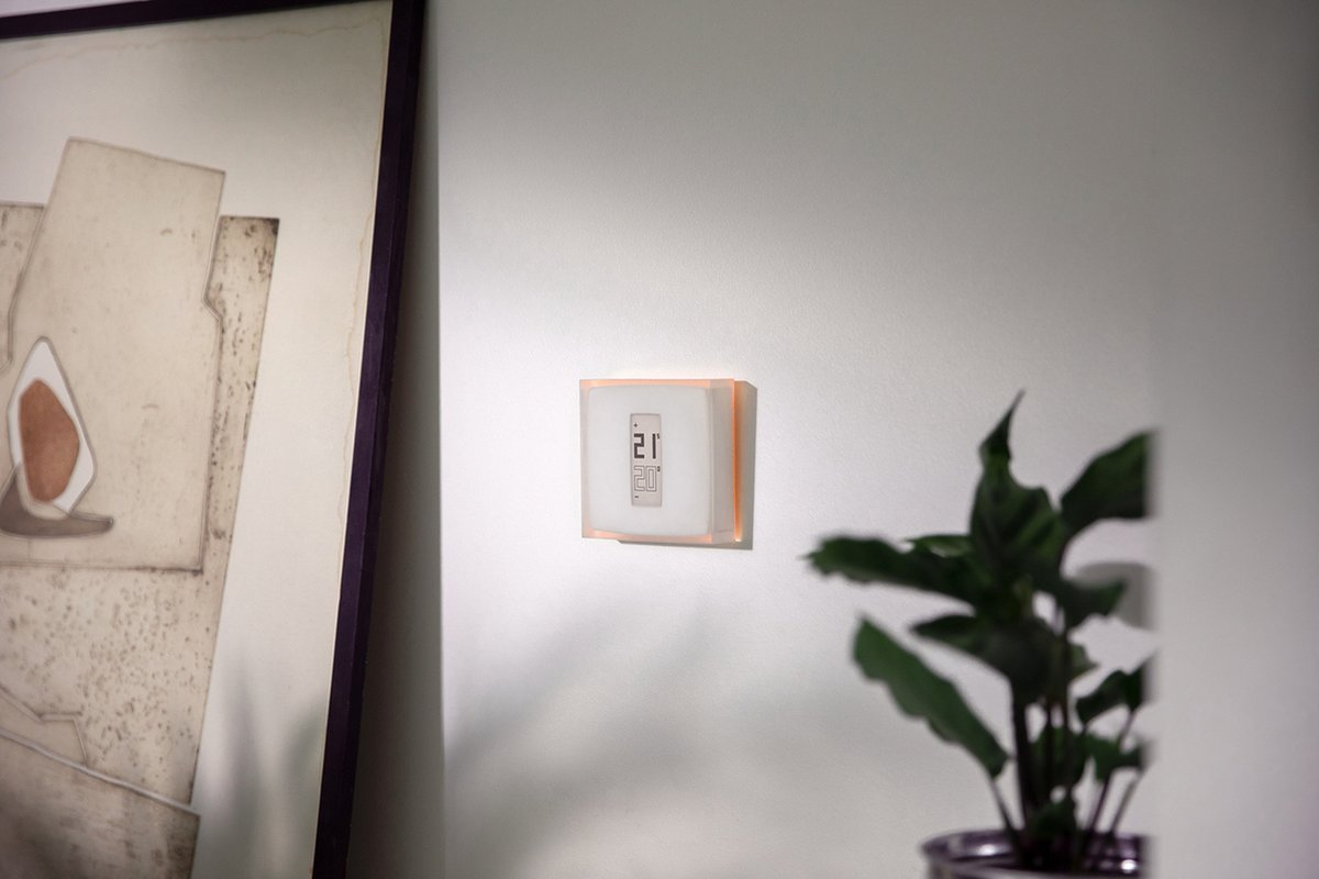 Un termostato blanco con pantalla digital brilla en naranja en una pared clara, junto a un cuadro enmarcado y una planta.