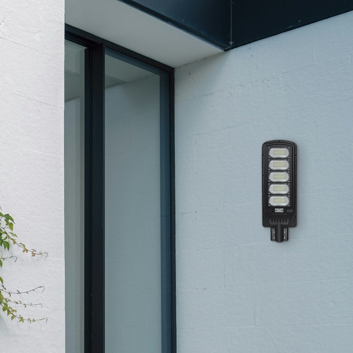 Una luce LED da esterno rettangolare nera è montata su un muro di mattoni azzurro vicino a un'alta finestra di vetro.