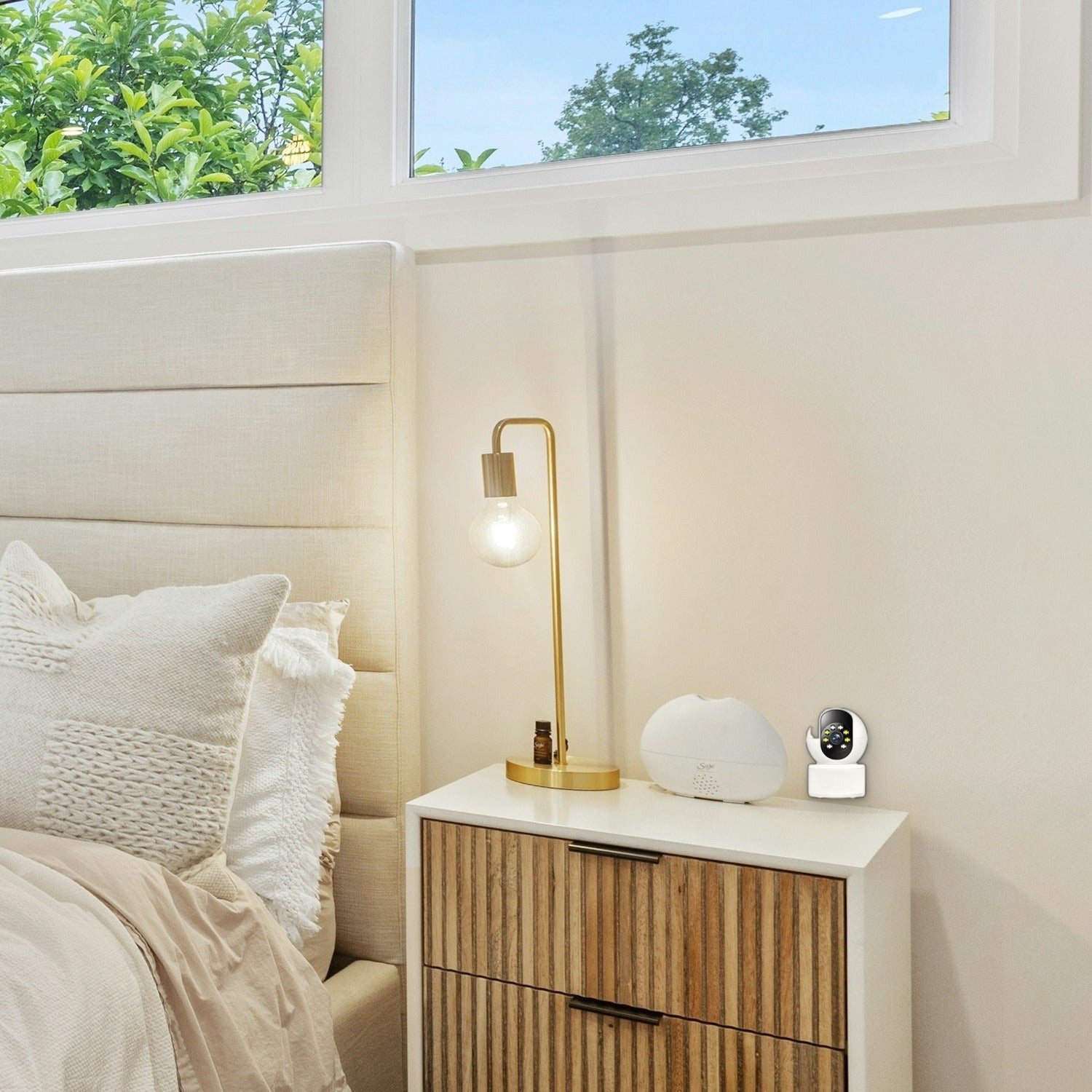 Una mesita de noche blanca con lámpara, difusor y cámara pequeña junto a una cama clara y una ventana.