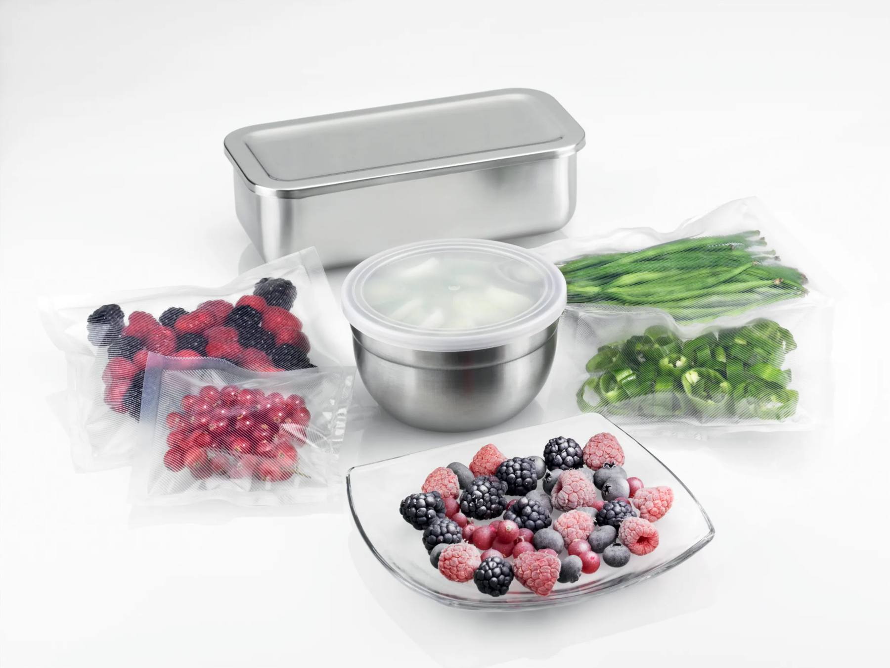 Edelstahlbehälter, Vakuumbeutel mit Beeren und Gemüse und ein Glasteller mit gefrorenen Beeren.