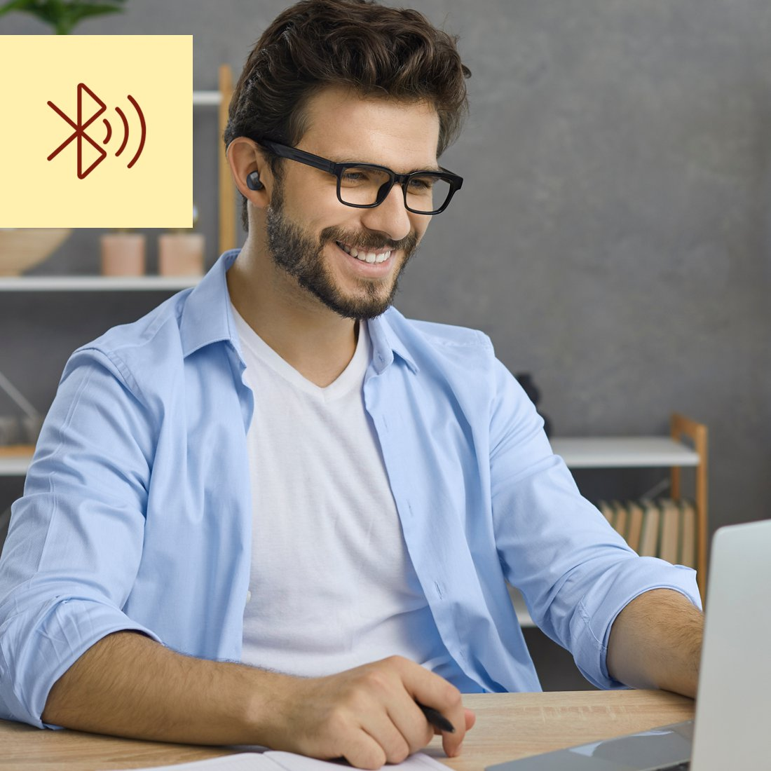 Ein lächelnder Mann mit Brille trägt drahtlose Ohrhörer und sitzt an einem Holztisch mit einem Laptop.