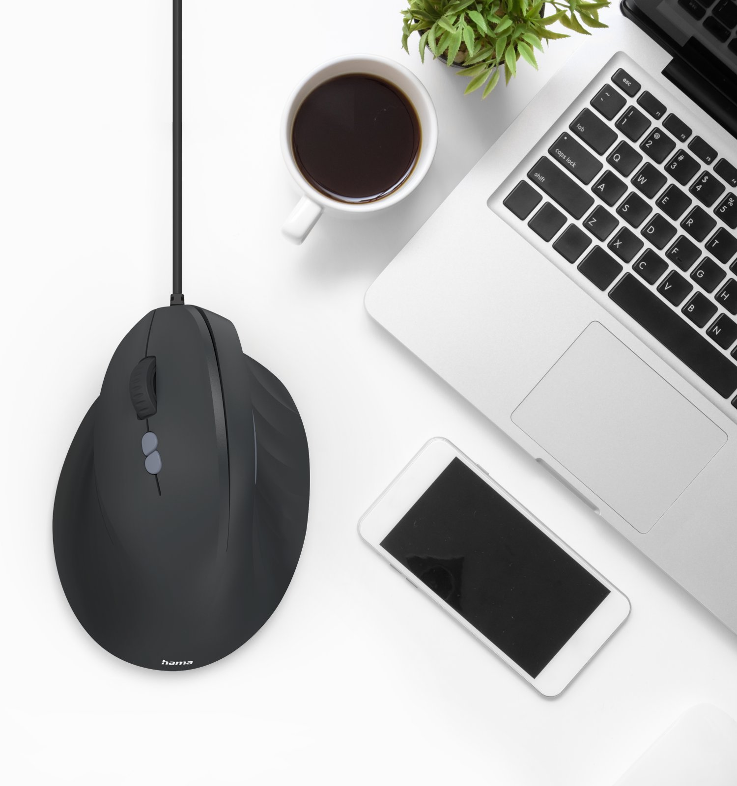 Vista cenital de un escritorio blanco con ratón negro con cable, ordenador portátil, móvil, taza y planta.
