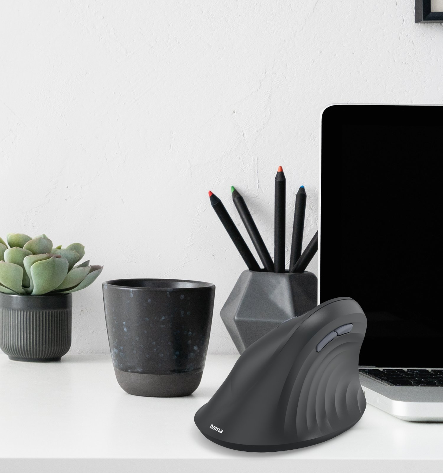 Un ratón de ordenador negro ergonómico en un escritorio junto a un portátil, bolígrafos y una planta en maceta.