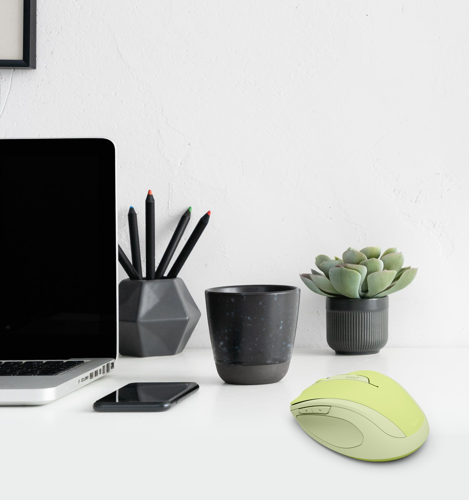 Una scrivania con laptop, tazza nera, matite, succulenta e mouse verde chiaro contro una parete bianca.