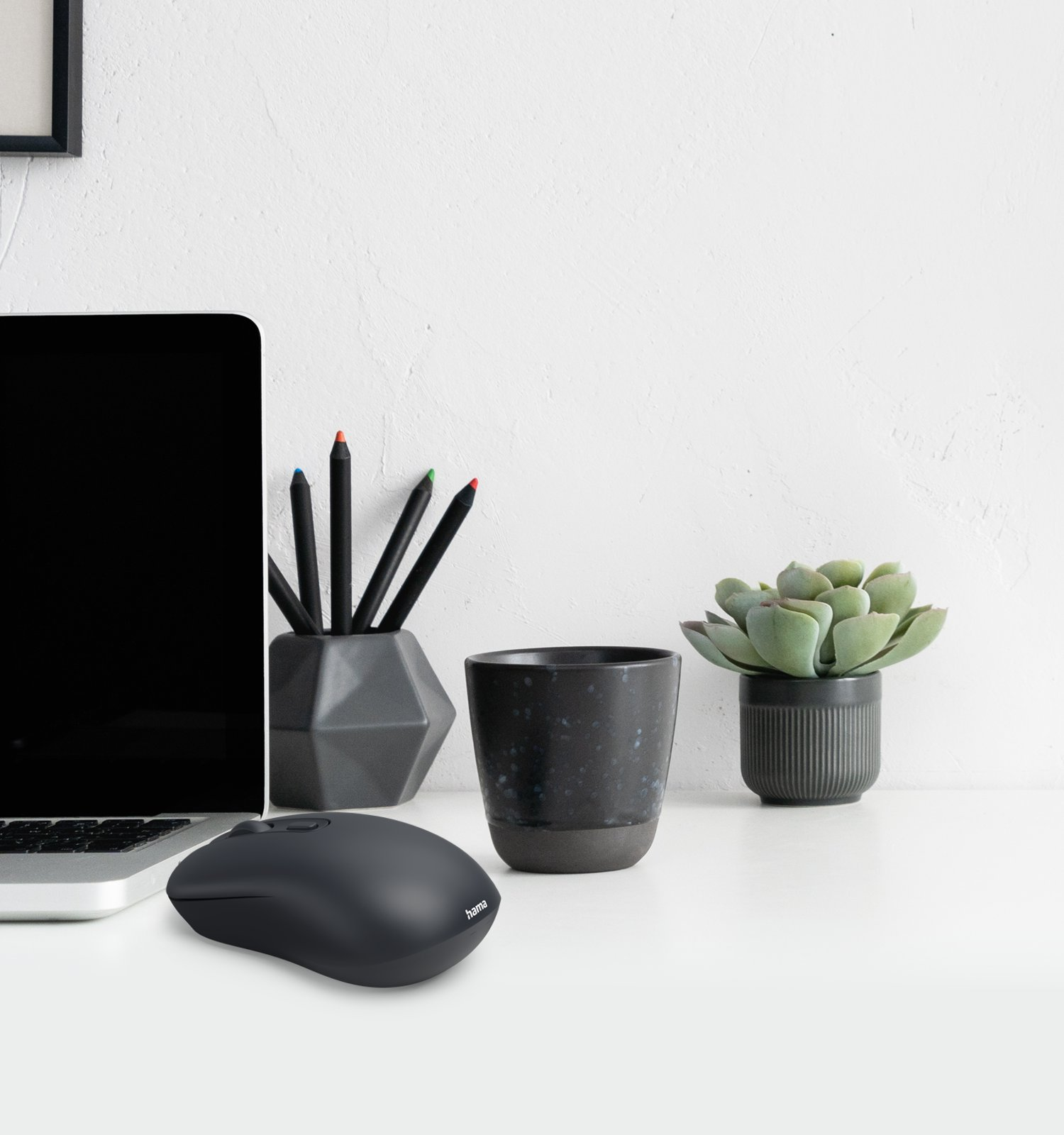 Un escritorio con laptop, ratón negro, portalápices, taza y una planta suculenta contra una pared blanca.
