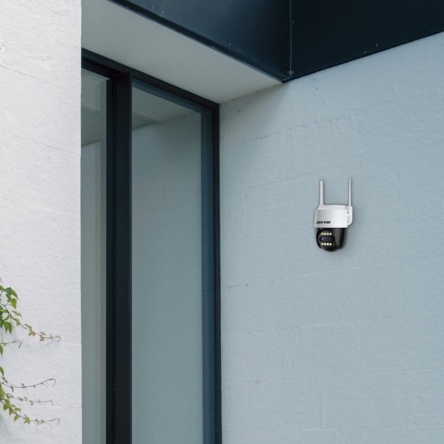 Cámara de seguridad blanca con dos antenas montada en una pared azul claro junto a una puerta de vidrio.