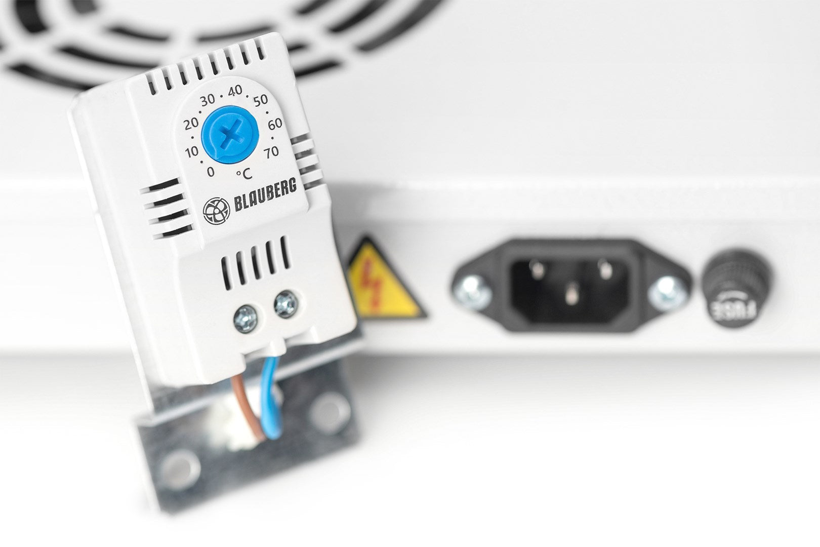 Ein weißer Blauberg-Thermostat mit blauem Einstellrad ist vor einem weißen Netzteil montiert.