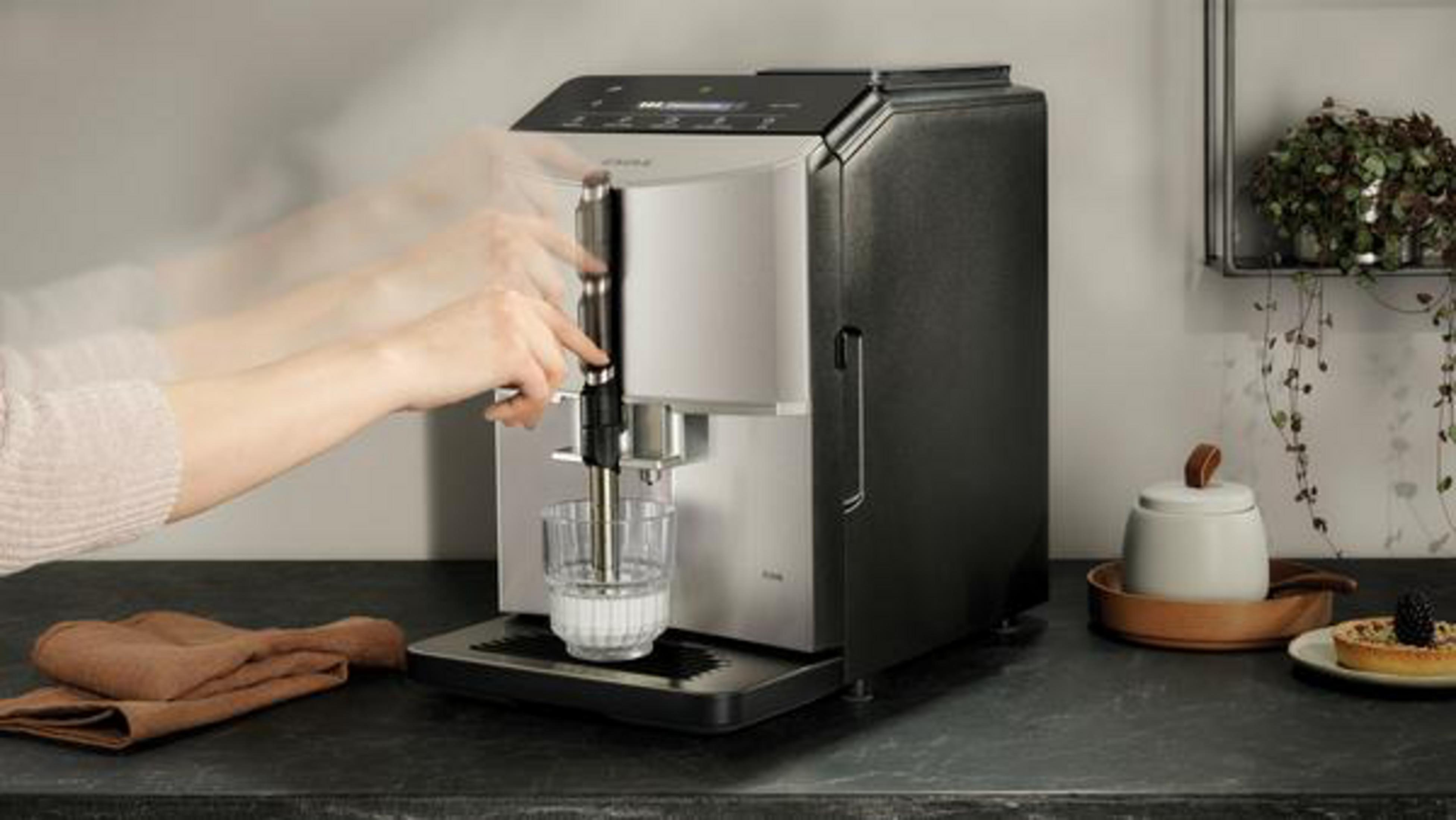 Eine Hand bewegt den Hebel einer silber-schwarzen Kaffeemaschine mit einer Glas Tasse mit Milch auf der Ablage.