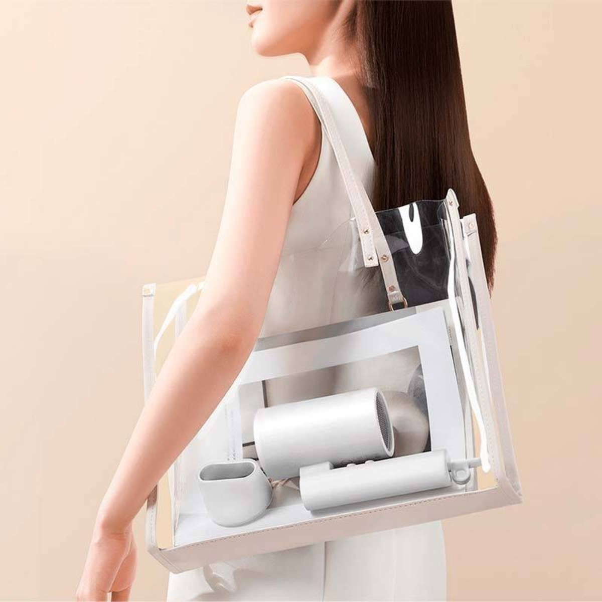 Una mujer lleva un bolso transparente que contiene un secador de pelo blanco, una revista y accesorios.