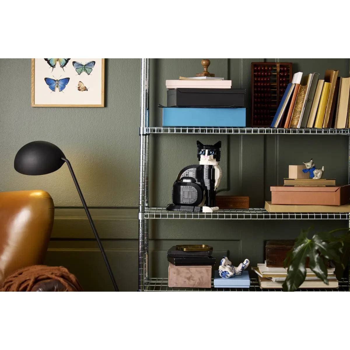 Una estantería metálica con libros y decoración se apoya en una pared verde junto a una lámpara de pie negra.