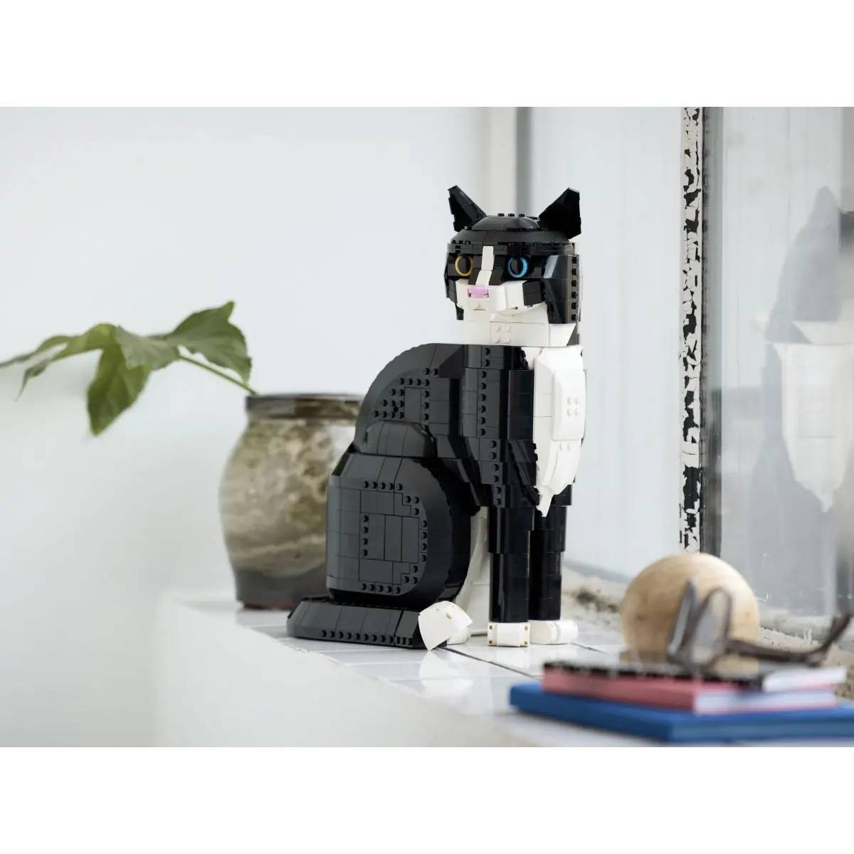 Una figura de Lego de un gato blanco y negro está sentada en el alféizar de una ventana junto a una planta.