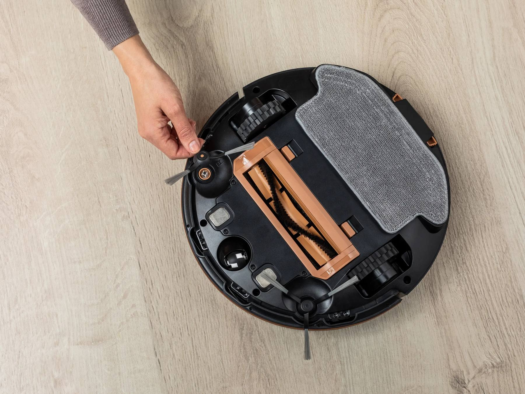 Una mano coloca el cepillo lateral de un robot aspirador negro en suelo de madera.