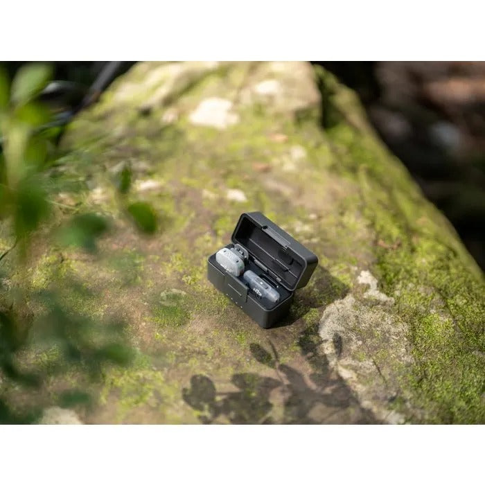 Estuche de carga negro abierto con micrófonos inalámbricos sobre una piedra con musgo al aire libre.