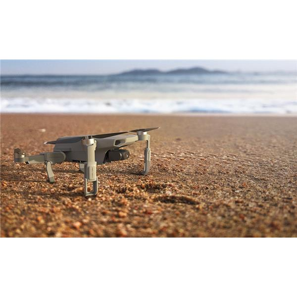 Eine kleine graue Drohne steht an einem Sandstrand mit dem Meer und fernen Hügeln im verschwommenen Hintergrund.