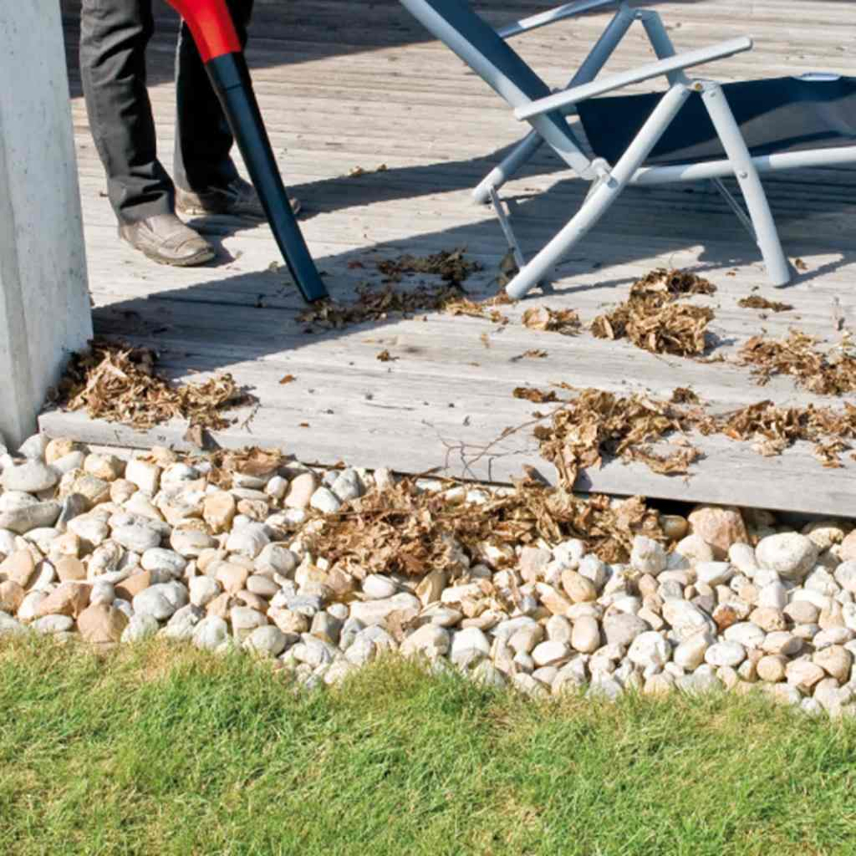 Person bläst trockene Blätter von einer Holzterrasse neben Kies und Rasen.
