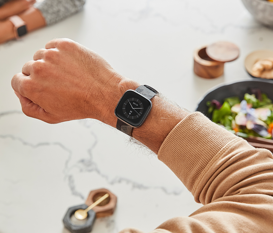 Arm mit schwarzer Smartwatch und gemustertem Armband auf weißem Tisch mit Salat.