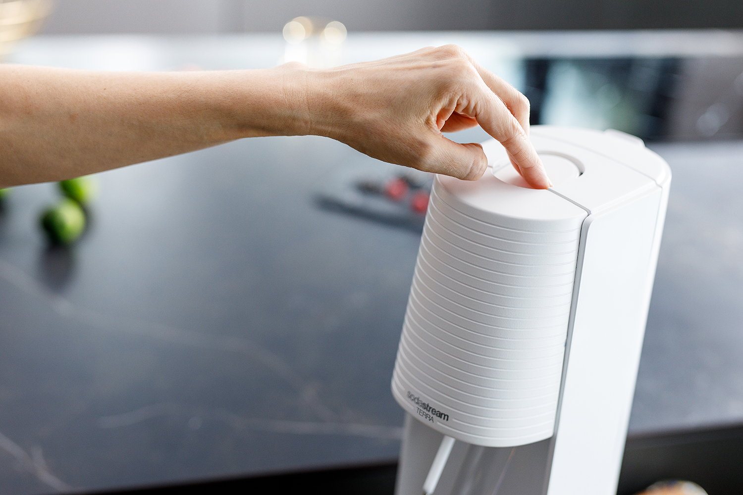 Eine Hand drückt den Knopf eines weißen Sodastream Terra Geräts auf einer dunklen Küchenarbeitsplatte.