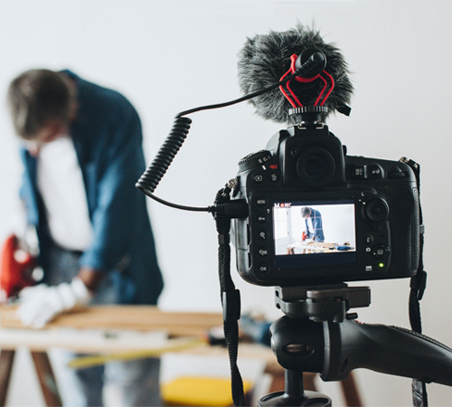 Eine DSLR-Kamera auf einem Stativ mit Mikrofon filmt einen Mann, der im Hintergrund an Holz arbeitet.