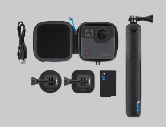 Una videocamera GoPro in custodia, cavo di ricarica, batteria, due supporti e un selfie stick nero su sfondo grigio.