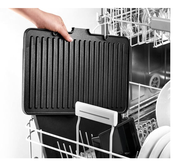 A hand places a black ribbed grill plate into the lower rack of an open dishwasher.