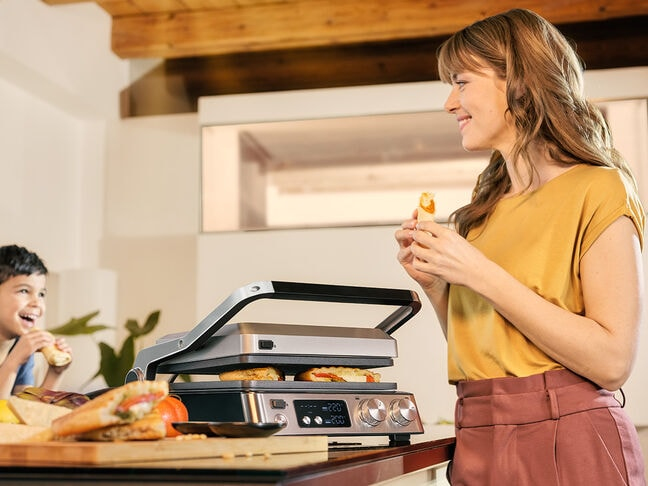 A woman holds food near a sandwich press on a counter, with a smiling child eating in the blurred background.