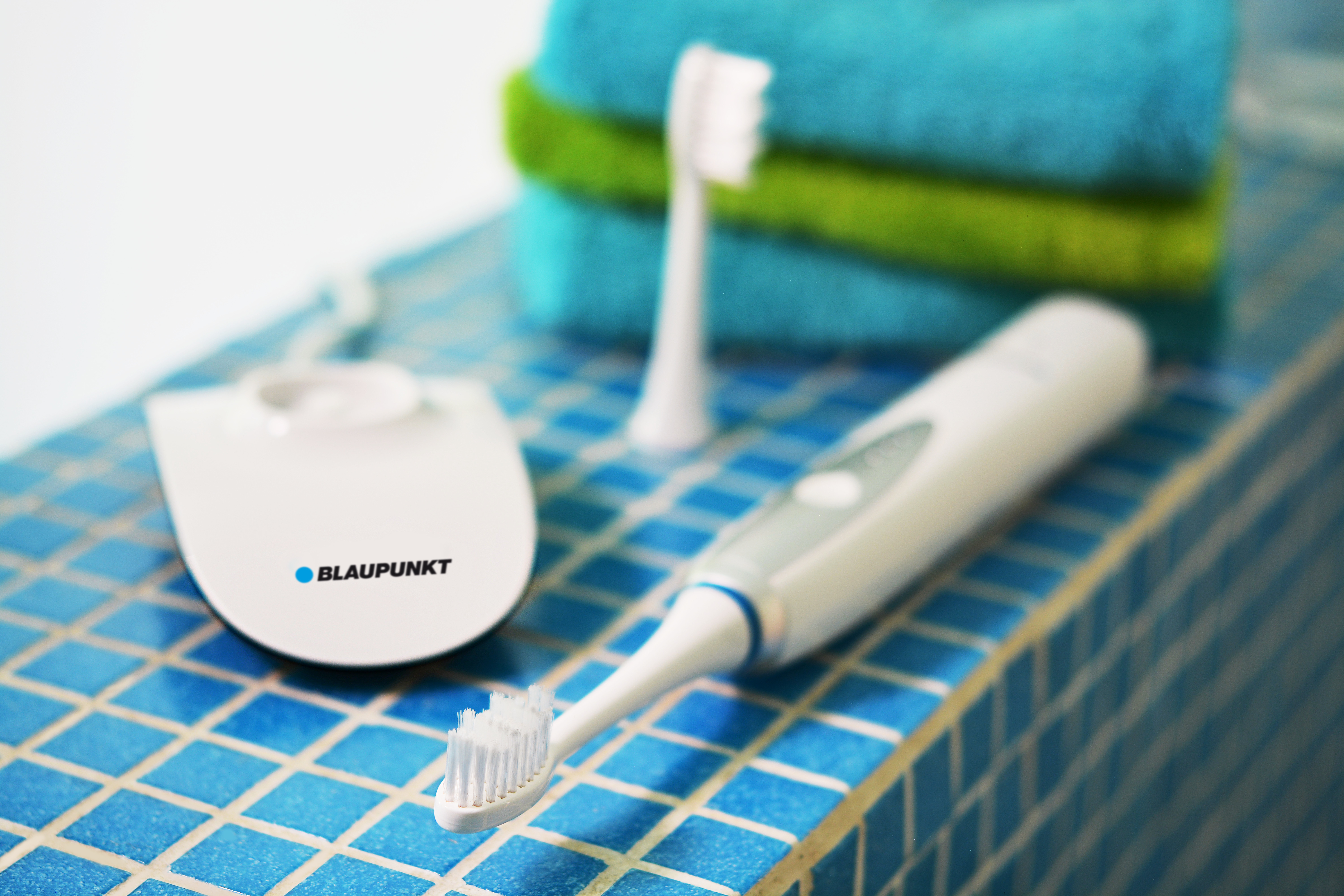 Uno spazzolino elettrico bianco Blaupunkt con caricatore su piastrelle blu, con asciugamani piegati sullo sfondo.