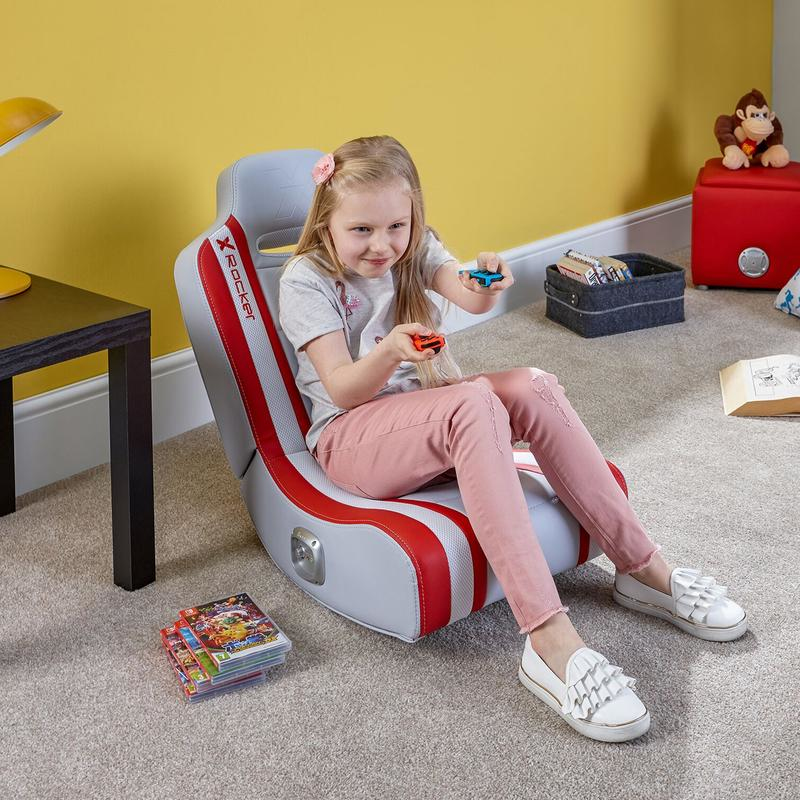 Ein junges Mädchen in pinken Hosen sitzt in einem grau-roten Gaming-Stuhl und hält Controller in einem hellen Zimmer.