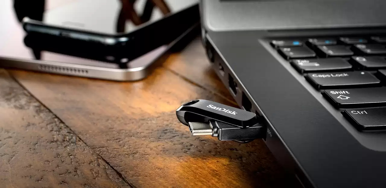 Ein schwarzer SanDisk-USB-Stick ist an einen Laptop auf einem Holztisch angeschlossen.