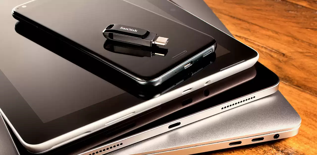 Ein SanDisk USB-Laufwerk liegt auf einem Smartphone, das auf gestapelten Tablets und Laptops auf einem Holztisch liegt.