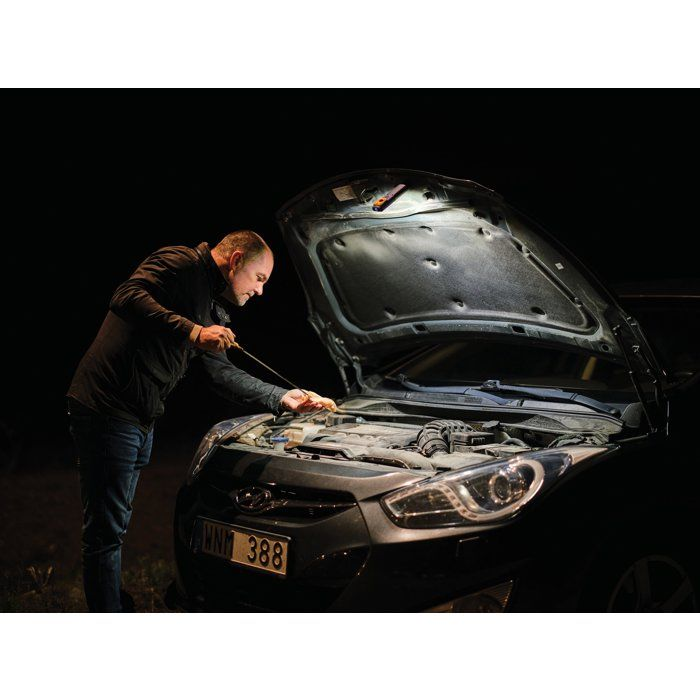 Ein Mann überprüft nachts bei geöffneter Motorhaube und künstlicher Beleuchtung den Ölstand seines Autos.