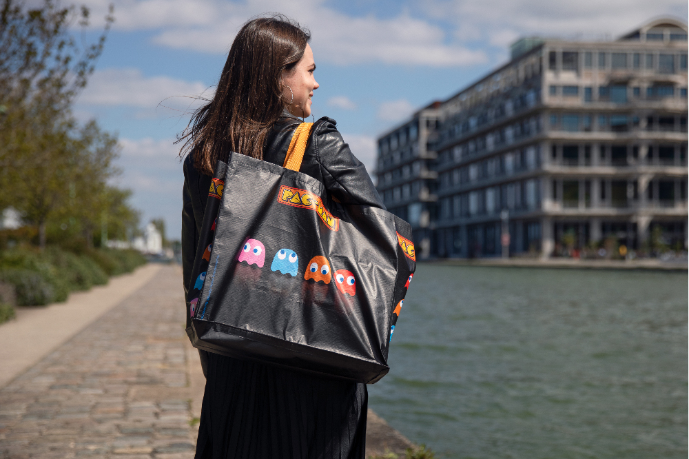 Eine Frau in schwarzer Lederjacke trägt eine große Pac-Man Tragetasche am Ufer eines Kanals.