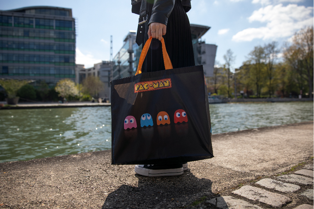 Eine Person hält eine schwarze Pac-Man-Stofftasche mit bunten Geistern an einem Flussufer und städtischen Gebäuden.