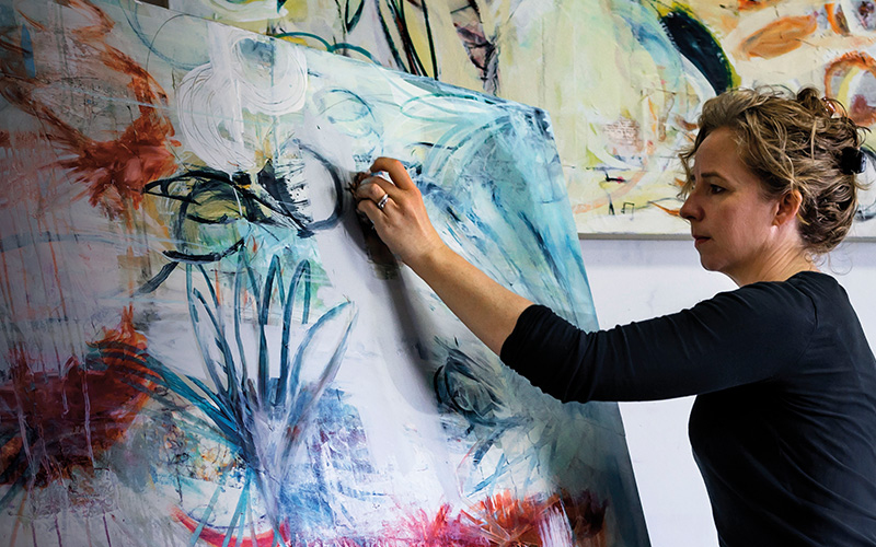 Un'artista in camicia nera dipinge una grande tela nel suo studio, applicando tratti astratti blu e rossi.
