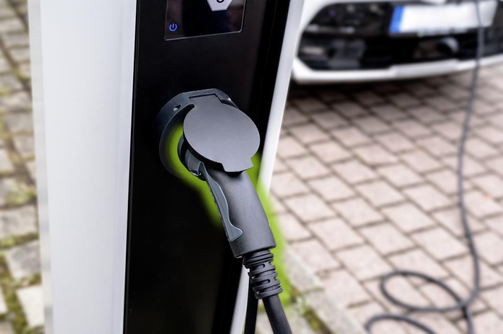 Bir elektrikli şarj kablosu bir şarj istasyonuna takılı, arka planda bir araba var.