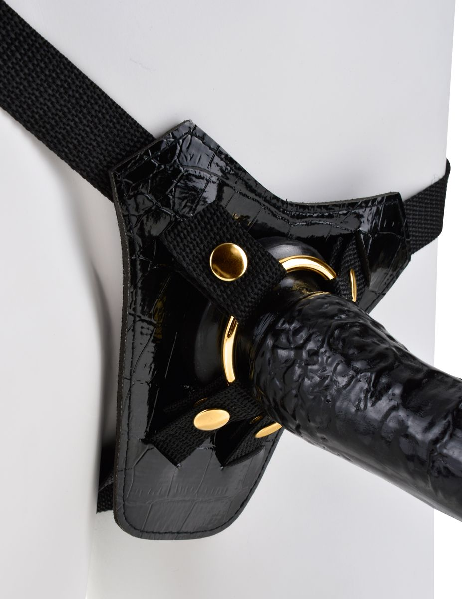 Ein schwarzer Ledergurt mit glänzendem Krokodilmuster, goldenen Akzenten und einem dicken, schwarzen Dildo.