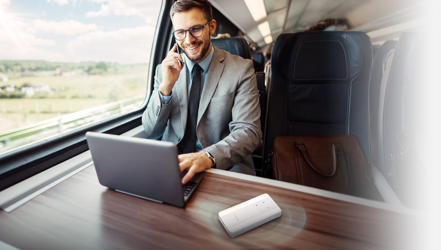 Een man in pak in een trein belt, gebruikt een laptop en een kleine router ligt op tafel.