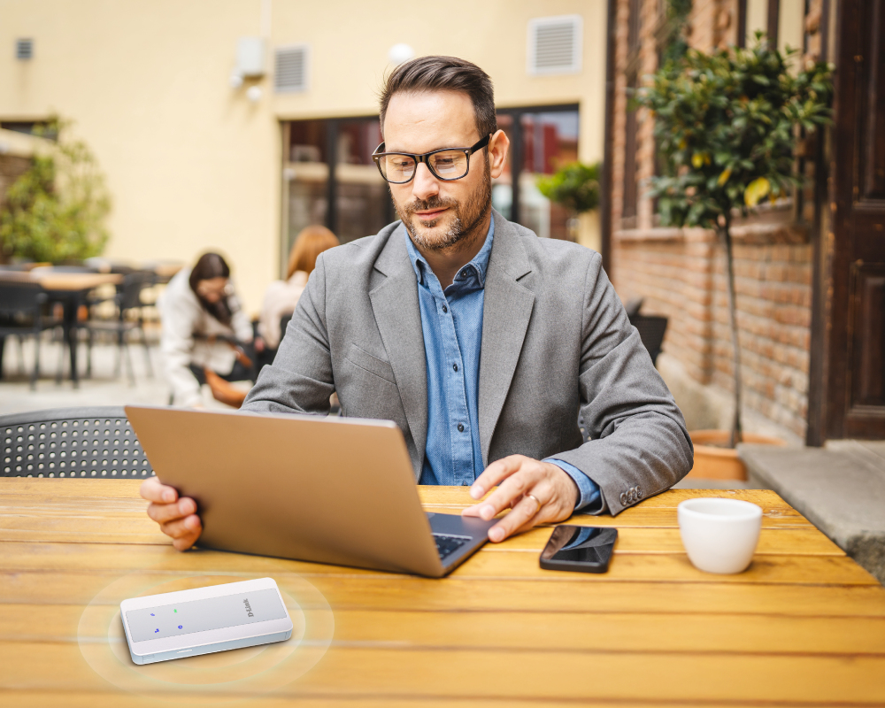 Een man met een bril werkt op een laptop in een café, met een D-Link-hotspot op de houten tafel.