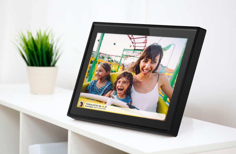 Ein schwarzer digitaler Bilderrahmen auf einem weißen Regal zeigt eine glückliche Familie bei einer Achterbahnfahrt.