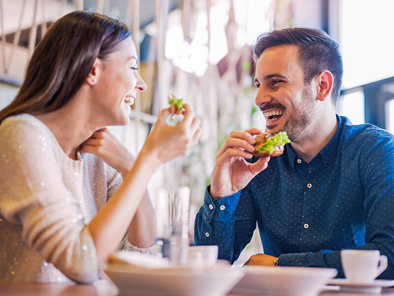 Una pareja joven riendo mientras come pequeñas hamburguesas en un café luminoso.