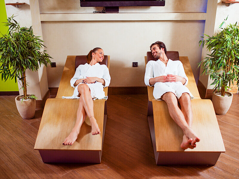 Una pareja feliz con albornoces blancos se relaja en tumbonas de madera en una zona de spa luminosa.