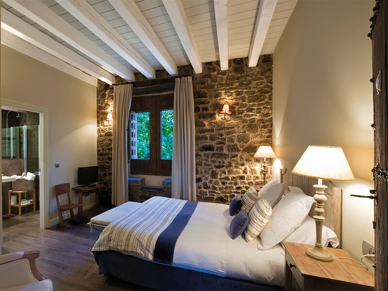 Acogedor dormitorio con pared de piedra, techo de vigas de madera blanca y cama grande.