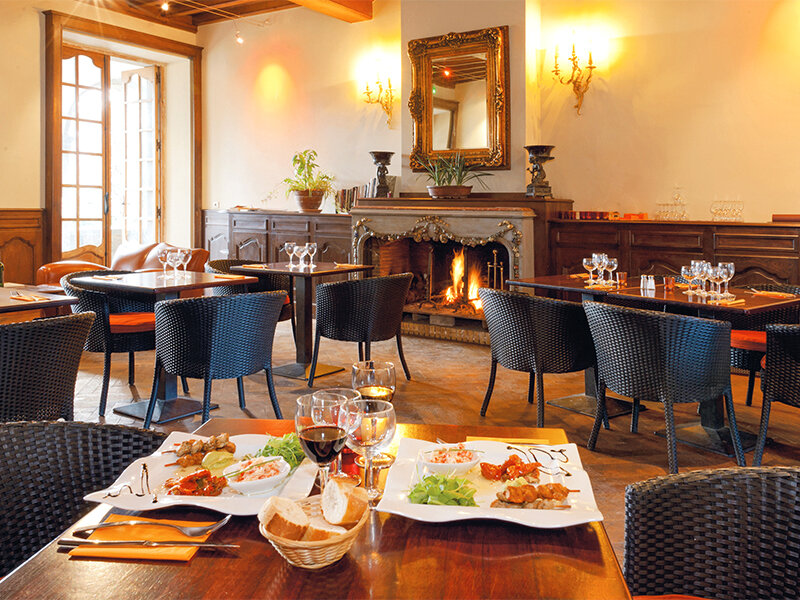 Cálido interior de restaurante con mesas de madera, sillas de mimbre negro, chimenea encendida y platos en primer plano.