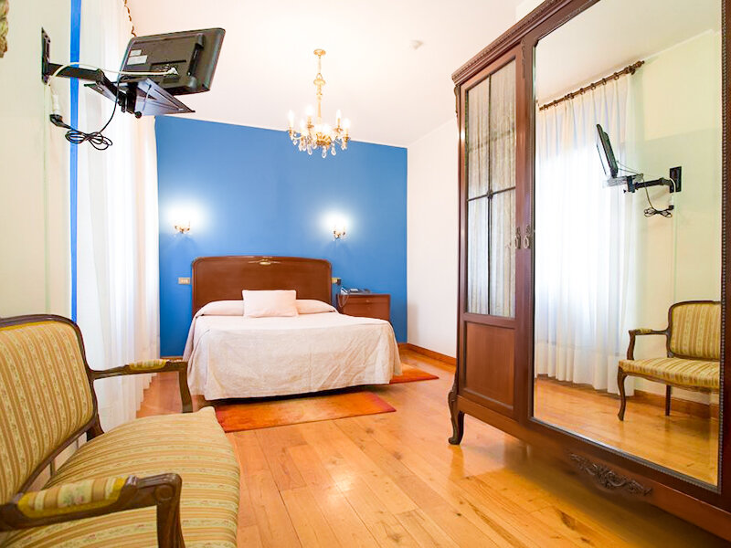 Dormitorio con suelo de madera, pared azul, cama doble, gran armario y sillones antiguos.