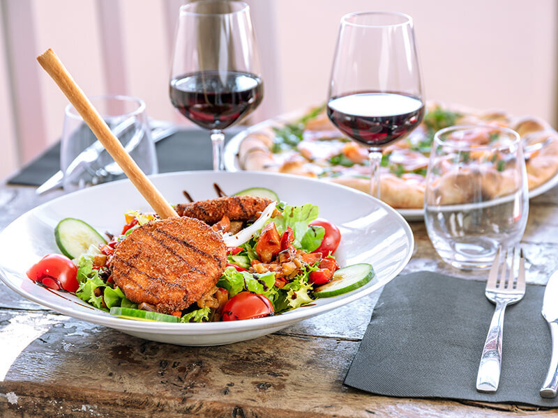 Un plato de ensalada fresca con hamburguesas empanadas, tomates, pepinos y colines, acompañado de vino tinto y pizza.