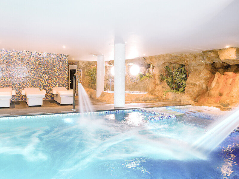 Una piscina cubierta con chorros, cascadas y tumbonas blancas frente a una pared de roca.