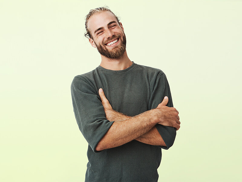 Un hombre sonriente con barba y el pelo recogido está de pie con los brazos cruzados sobre un fondo claro.